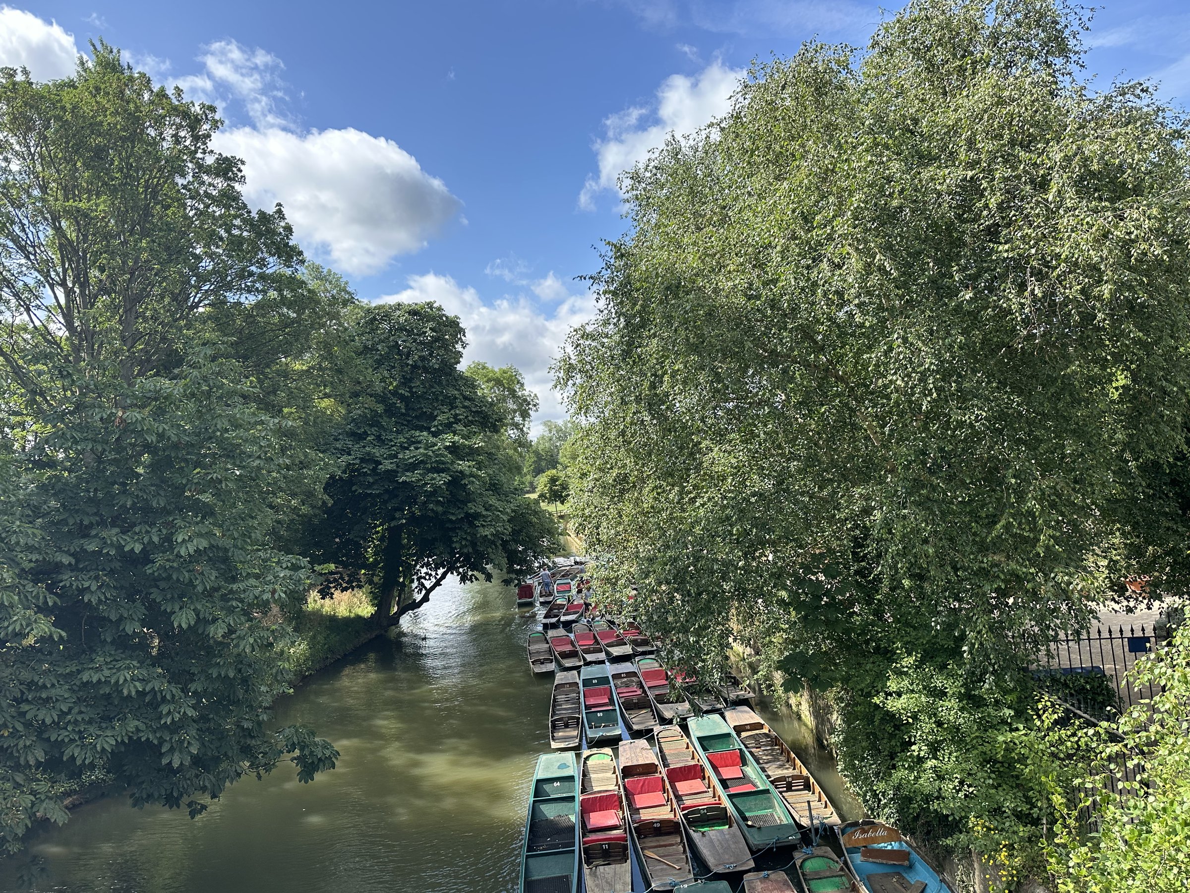 Punts at rest on the Cherwell, framed by tall trees and an open sky.