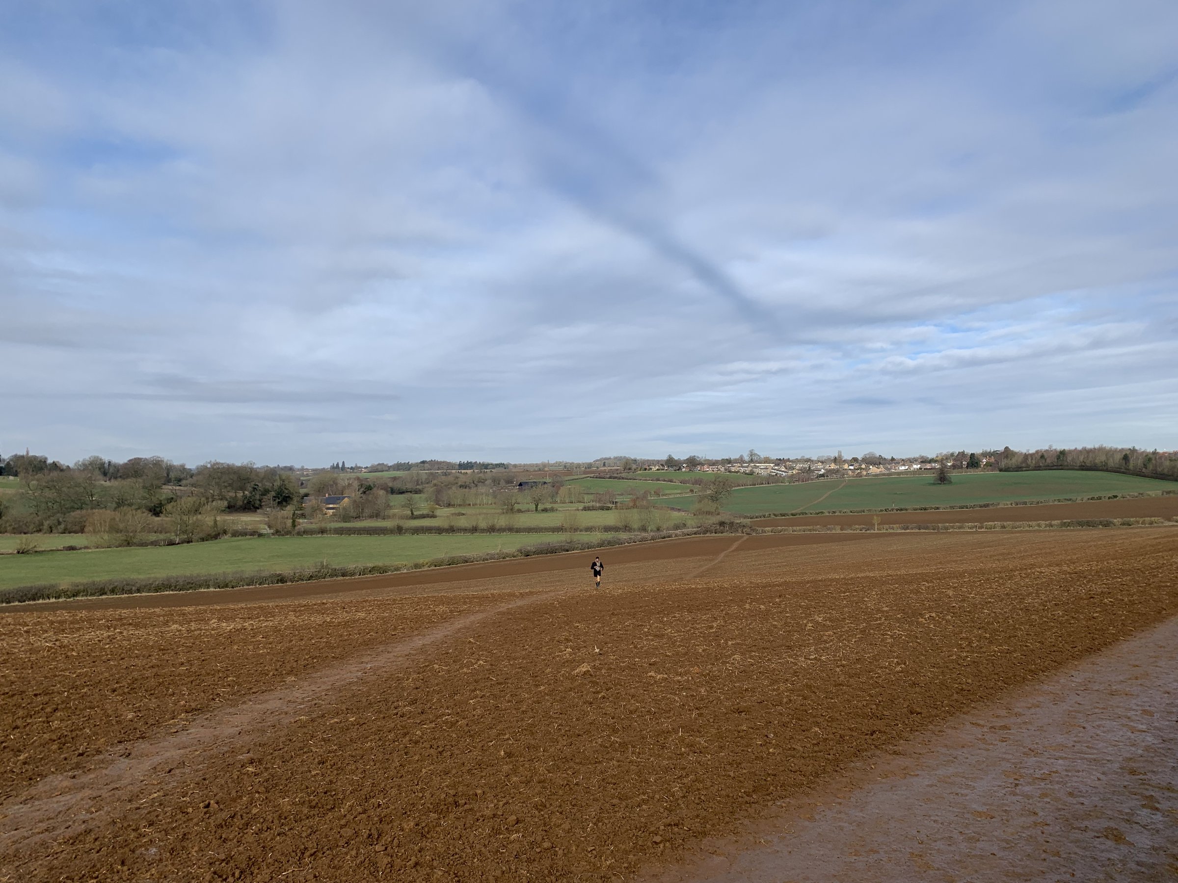 A vast ploughed field stretching to a low horizon under a wide cloud-streaked sky, a small figure walking in the middle distance.